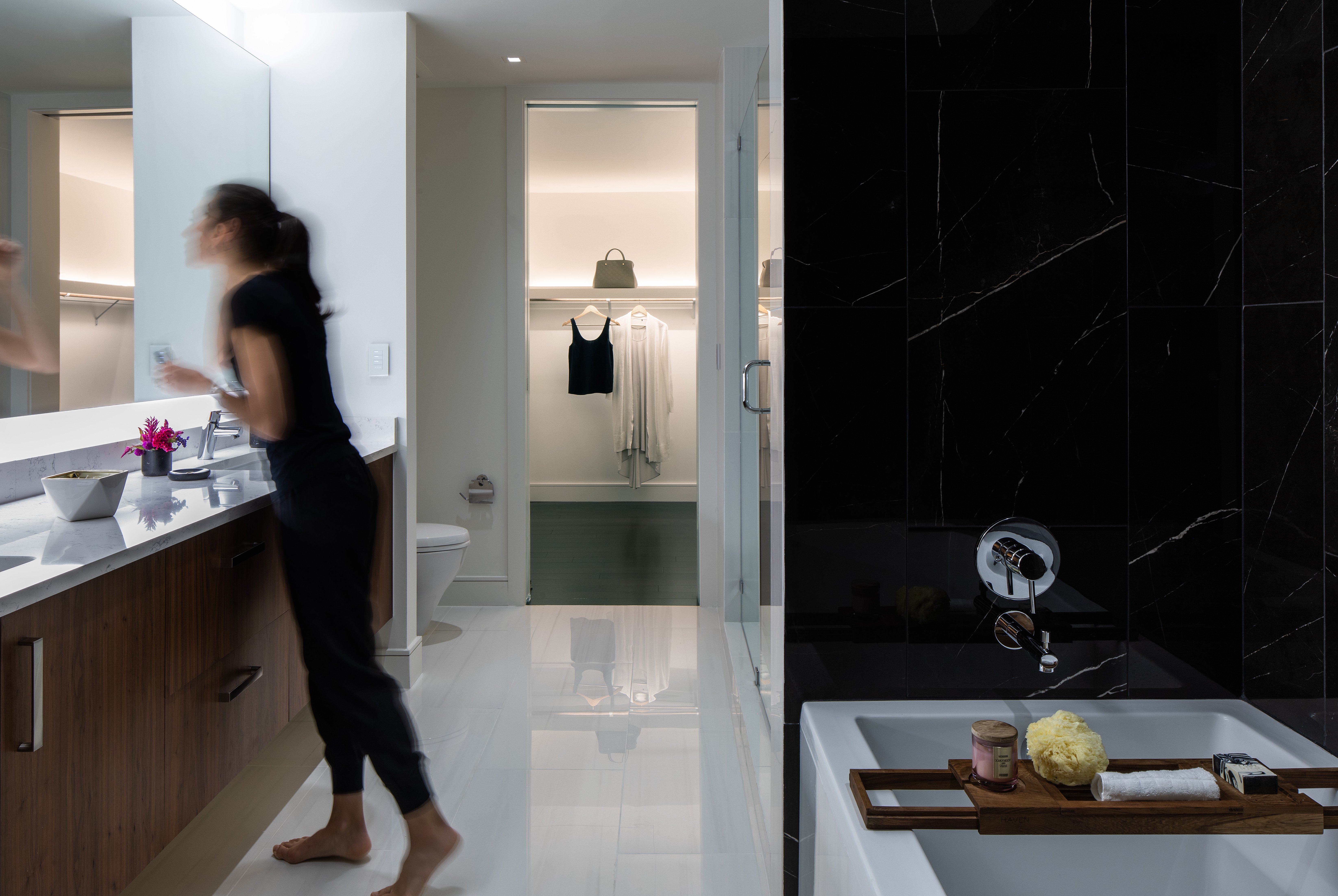 A woman leaning into a mirror in a bathroom brightly lit with downlights and coves at a cool, white light.
