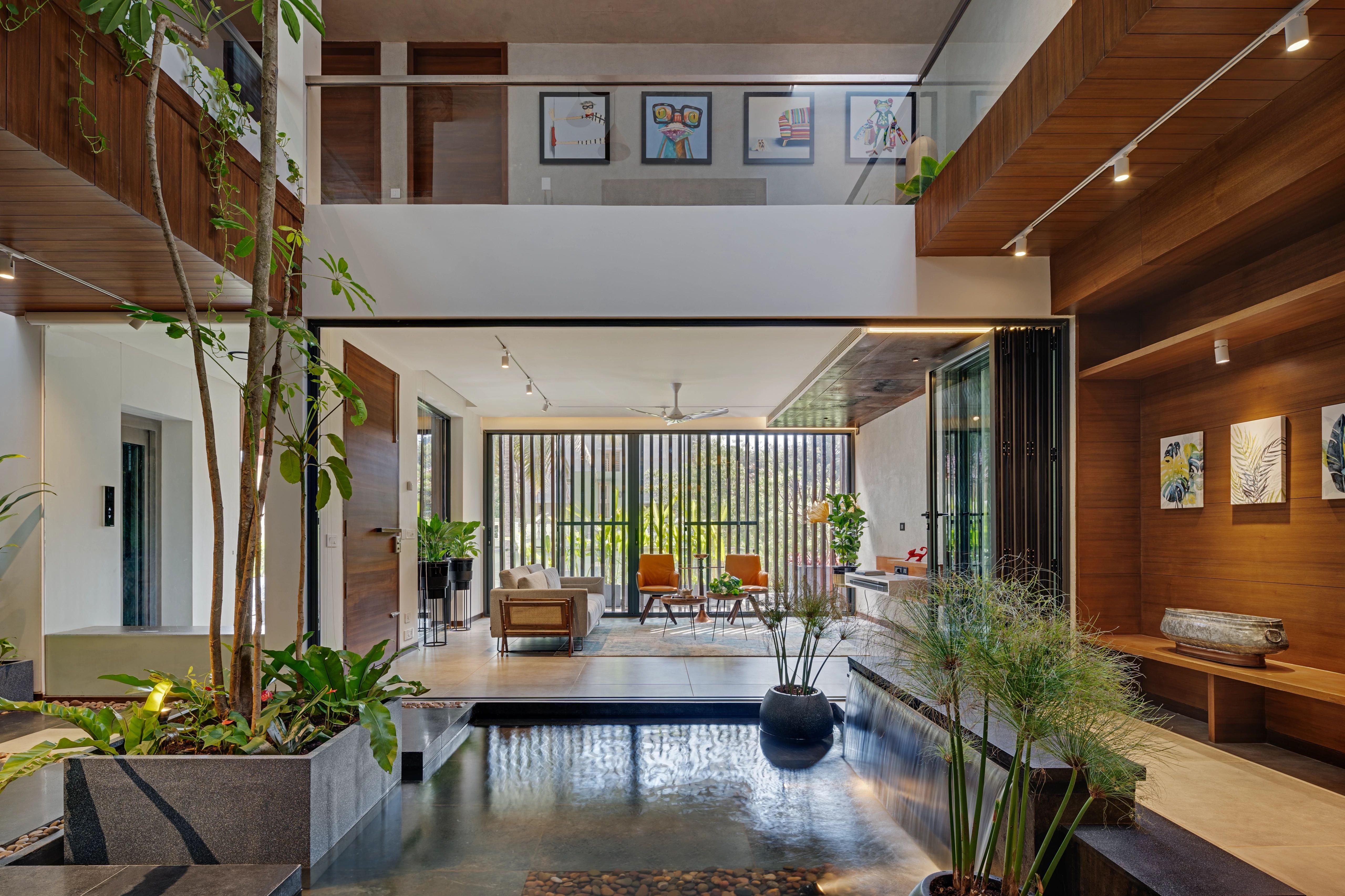 The interior of a house with a green-space with plants and a fountain next to a lounge area with a lot of daylight streaming in. 